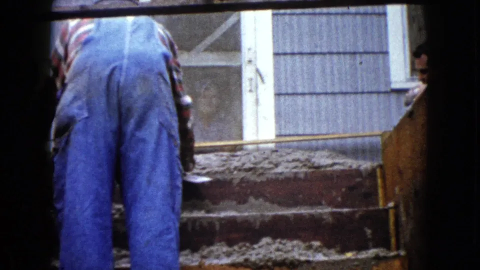 1960 home-movie frame from Wauconda showing a worker from behind as wet cement covers several wooden stair forms.