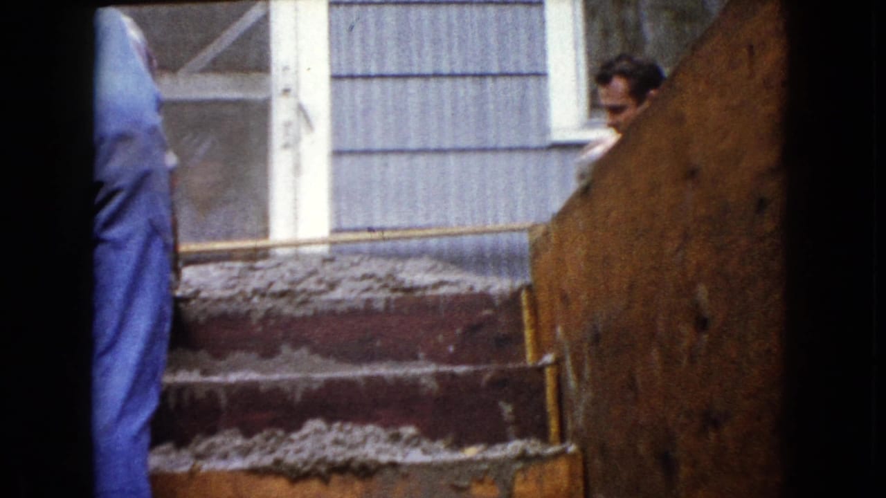 1960 Wauconda home-movie frame showing men working near fresh concrete steps outside a residential doorway and siding.