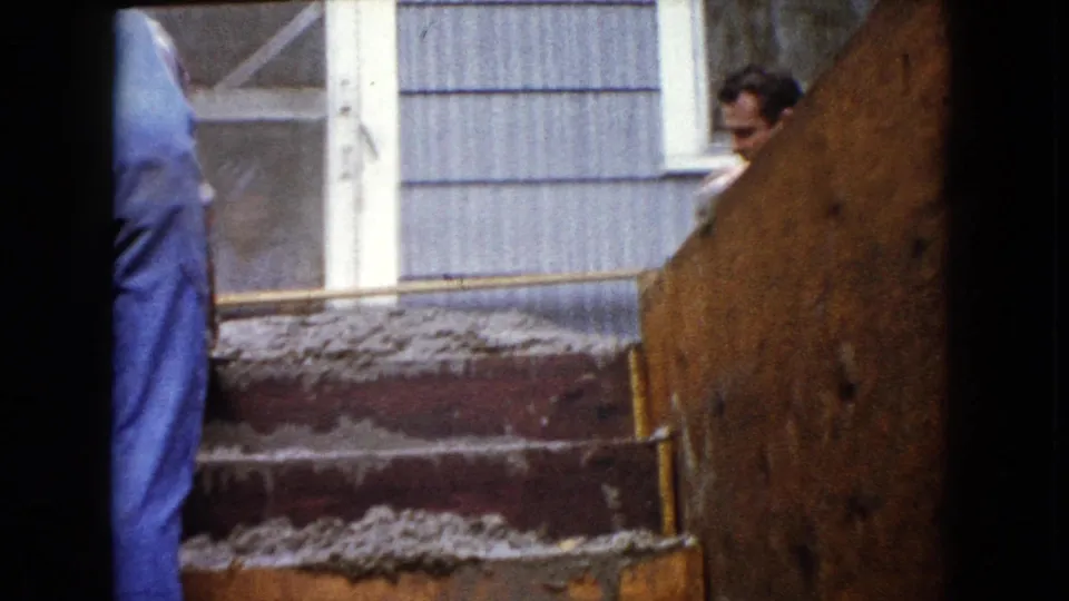 1960 Wauconda home-movie frame showing men working near fresh concrete steps outside a residential doorway and siding.