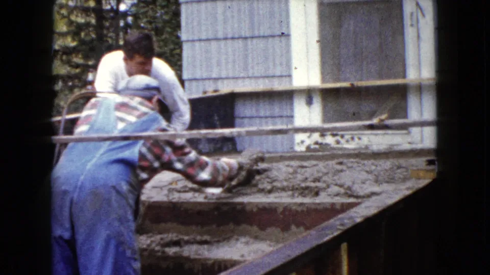 A man in blue overalls uses a trowel on wet concrete stairs as another man assists outside a Wauconda residence.