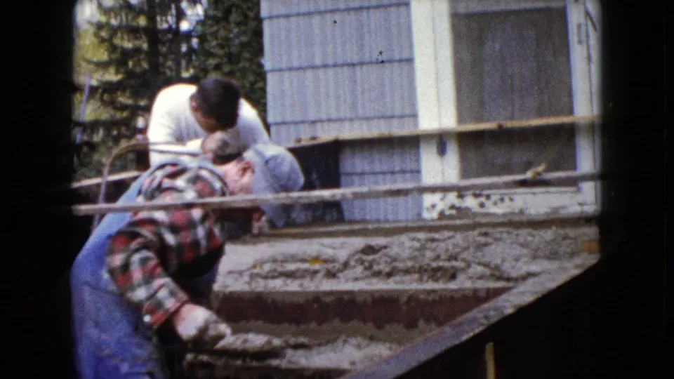 Two men continue smoothing a fresh concrete stair surface in 1960 Wauconda residential home-movie footage.