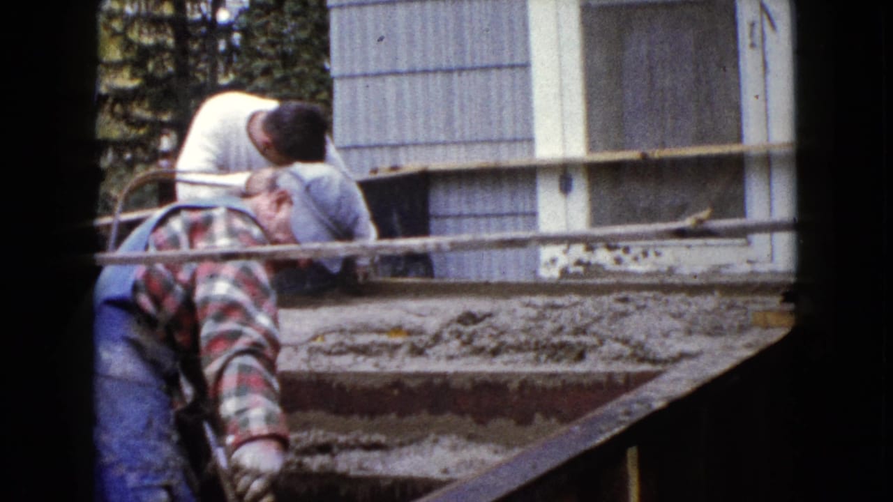 In 1960 Wauconda, Illinois footage, two adult men lean over wet cement steps beside a light-sided house.