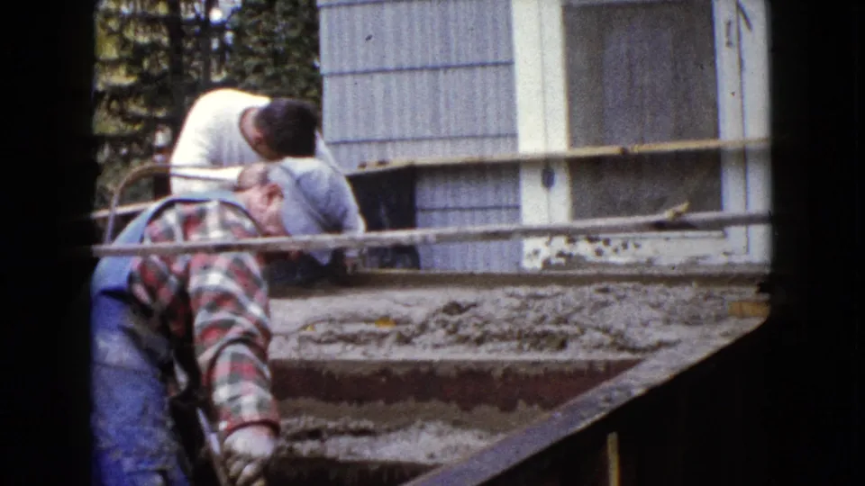 In 1960 Wauconda, Illinois footage, two adult men lean over wet cement steps beside a light-sided house.