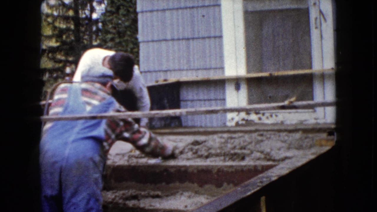 A man in blue overalls uses a trowel on wet cement steps while another adult assists beside the Wauconda house exterior.