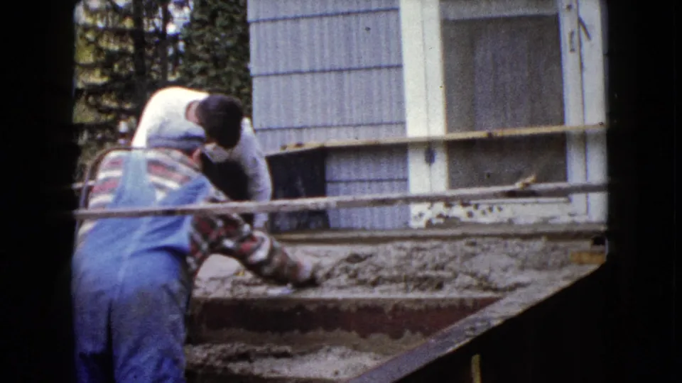 A man in blue overalls uses a trowel on wet cement steps while another adult assists beside the Wauconda house exterior.