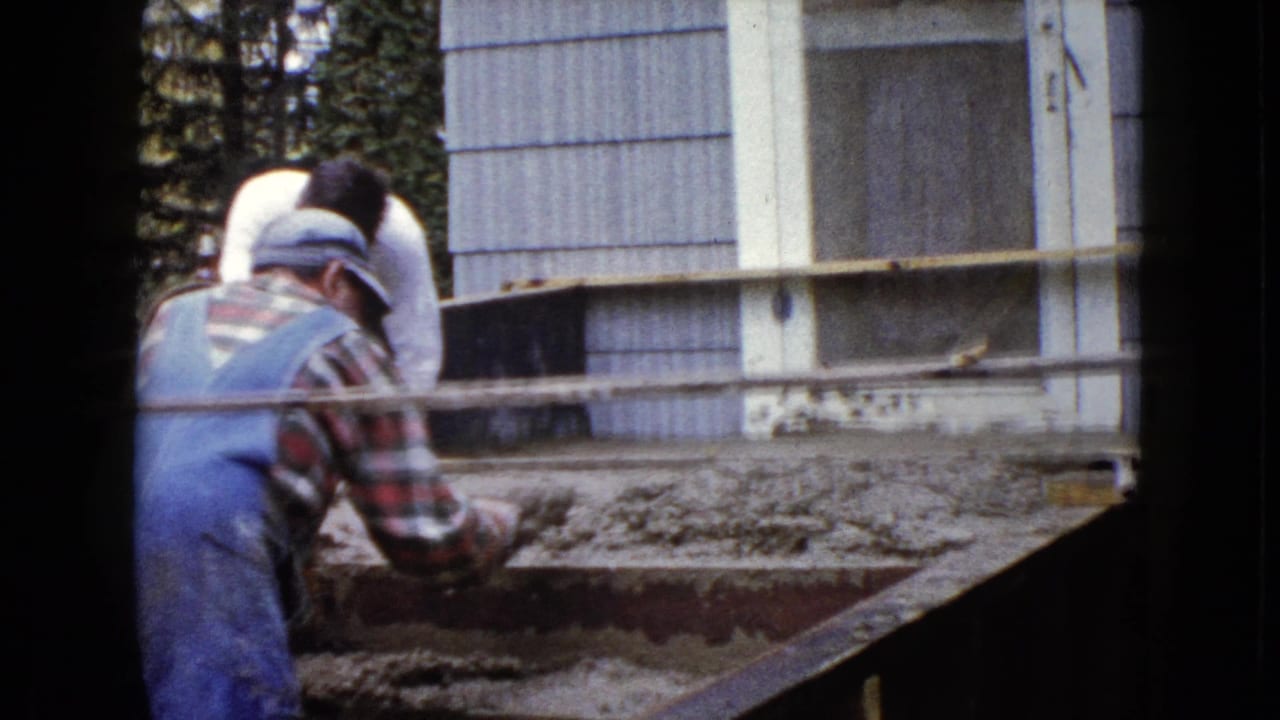 The overalled worker continues smoothing wet cement on exterior steps, with house siding and windows filling the background.