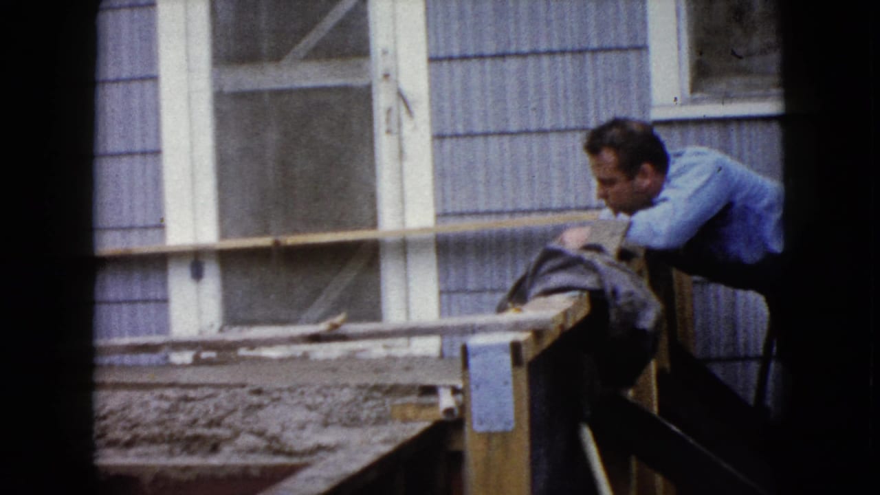 Man in a blue shirt leans over wooden forms with wet concrete beside a house window.