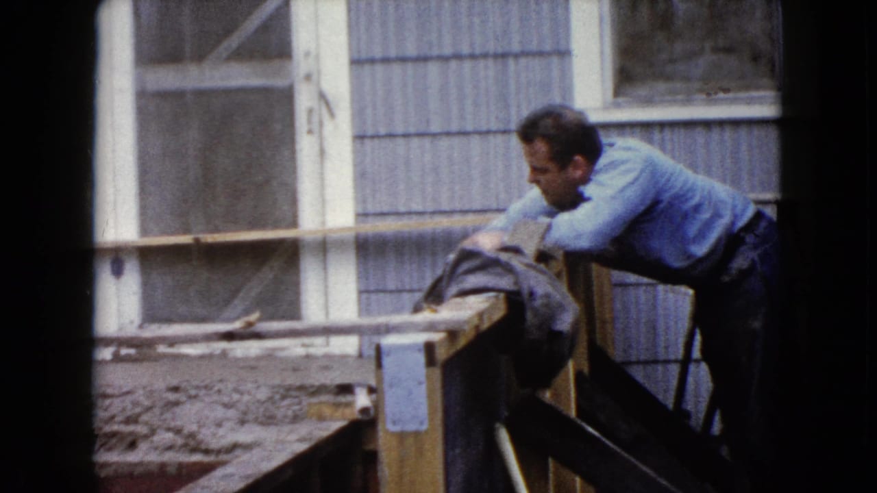 Man in a blue shirt bends over a wooden form while working concrete near a house wall.