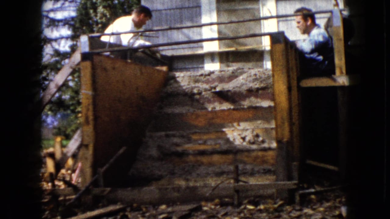 Two men are visible at a residential concrete step project, with wooden forms, railing, siding, and yard debris in view.