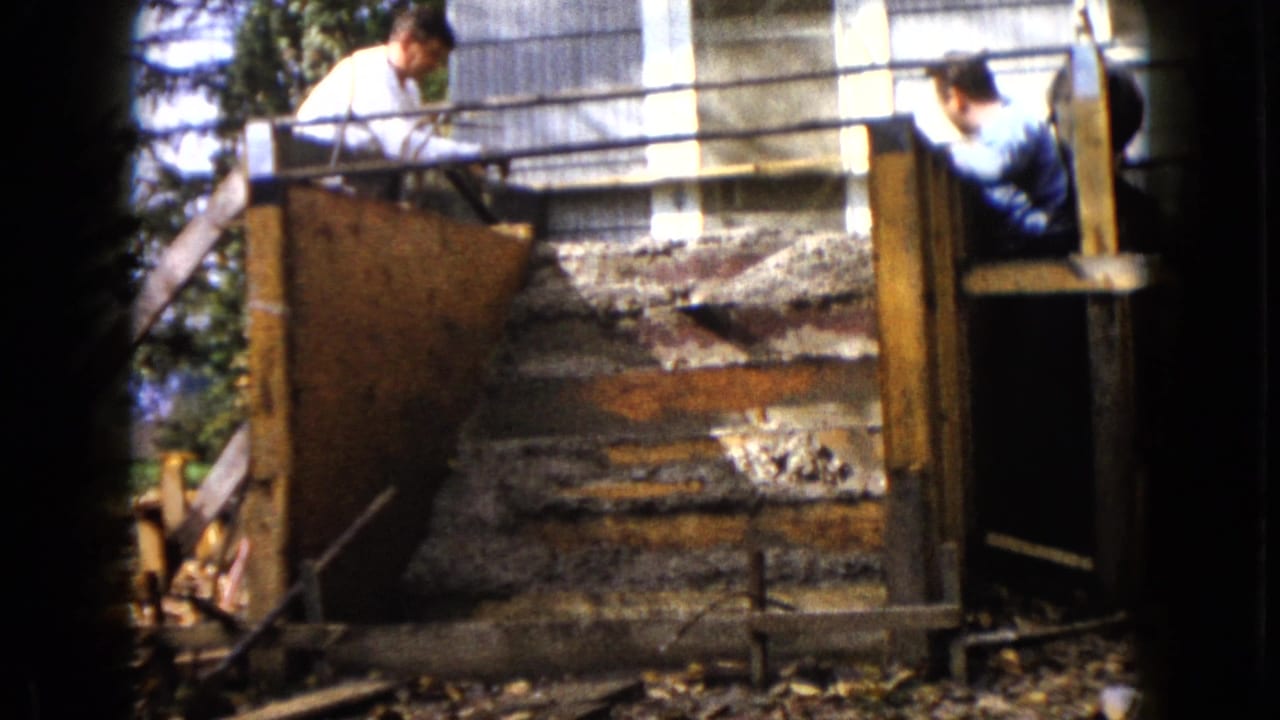 In Wauconda, Illinois, two men appear beside wooden forms and rough concrete steps during a 1960 8mm home-movie construction scene.