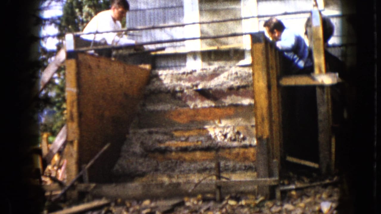 Two men work over wooden forms and concrete steps beside a building exterior in this 1960 Wauconda home-movie frame.