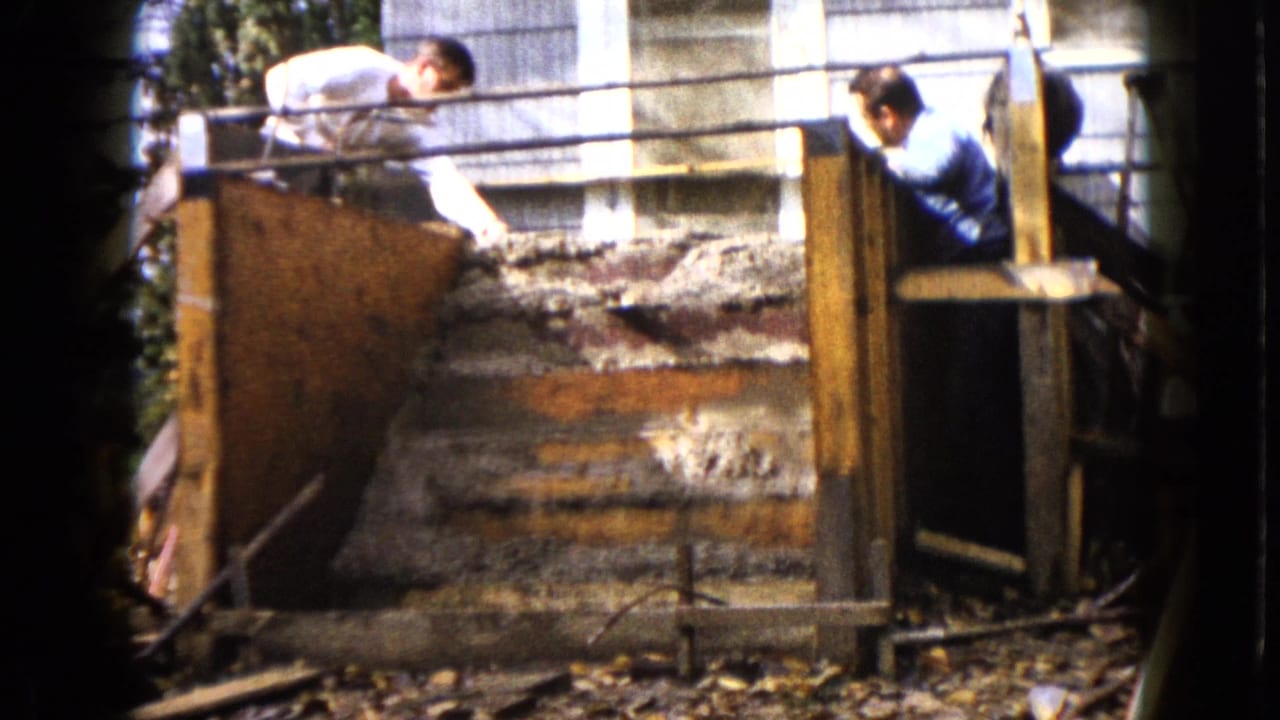 A man in a white shirt reaches down toward wet concrete steps while another man in blue stands near the form in Wauconda, Illinois, 1960.
