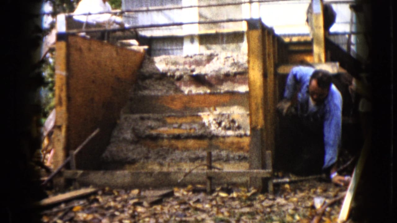 Man in a blue shirt crouches beside a wooden frame while another man works above.