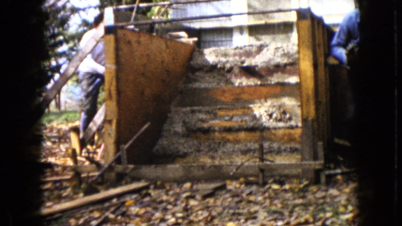 The camera frames the wooden structure from the yard as a man in a white shirt stands to the left, with another worker partly visible at the edge.