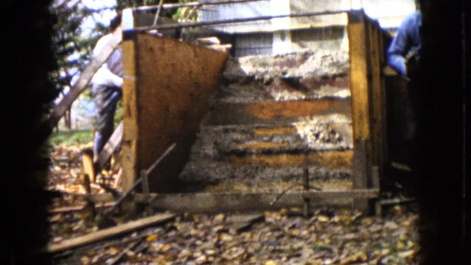 The camera frames the wooden structure from the yard as a man in a white shirt stands to the left, with another worker partly visible at the edge.
