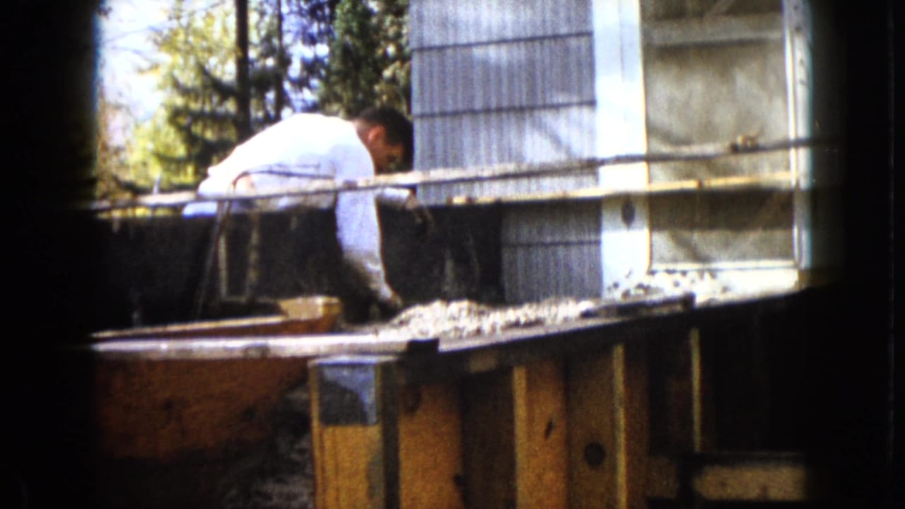 A man bends over the upper edge of the backyard wooden frame, with corrugated siding and lumber visible in the 1960 Wauconda footage.