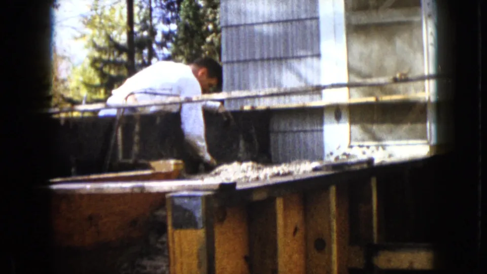 A man bends over the upper edge of the backyard wooden frame, with corrugated siding and lumber visible in the 1960 Wauconda footage.