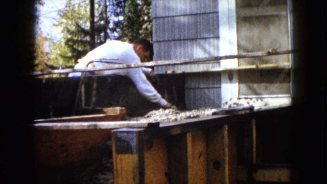 In Wauconda, Illinois in 1960, a man bends over a wooden plank or work surface near house siding and trees.