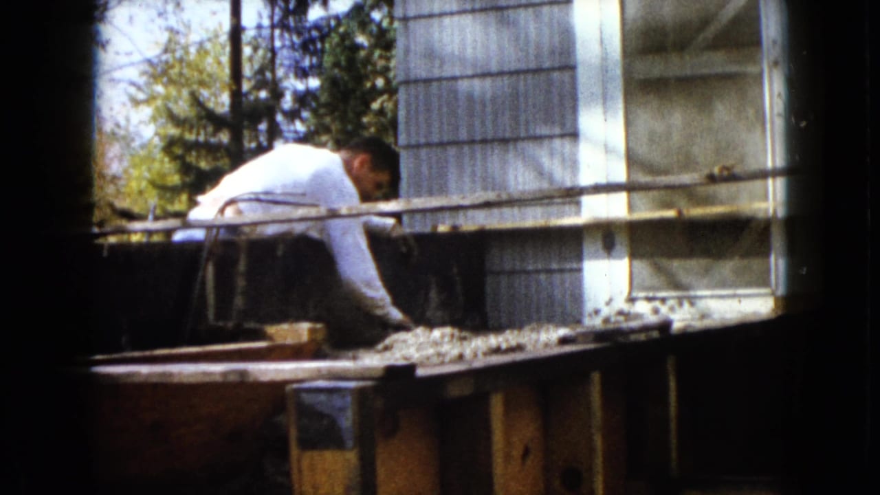 Man continues working over a debris-covered wooden plank near a house wall.