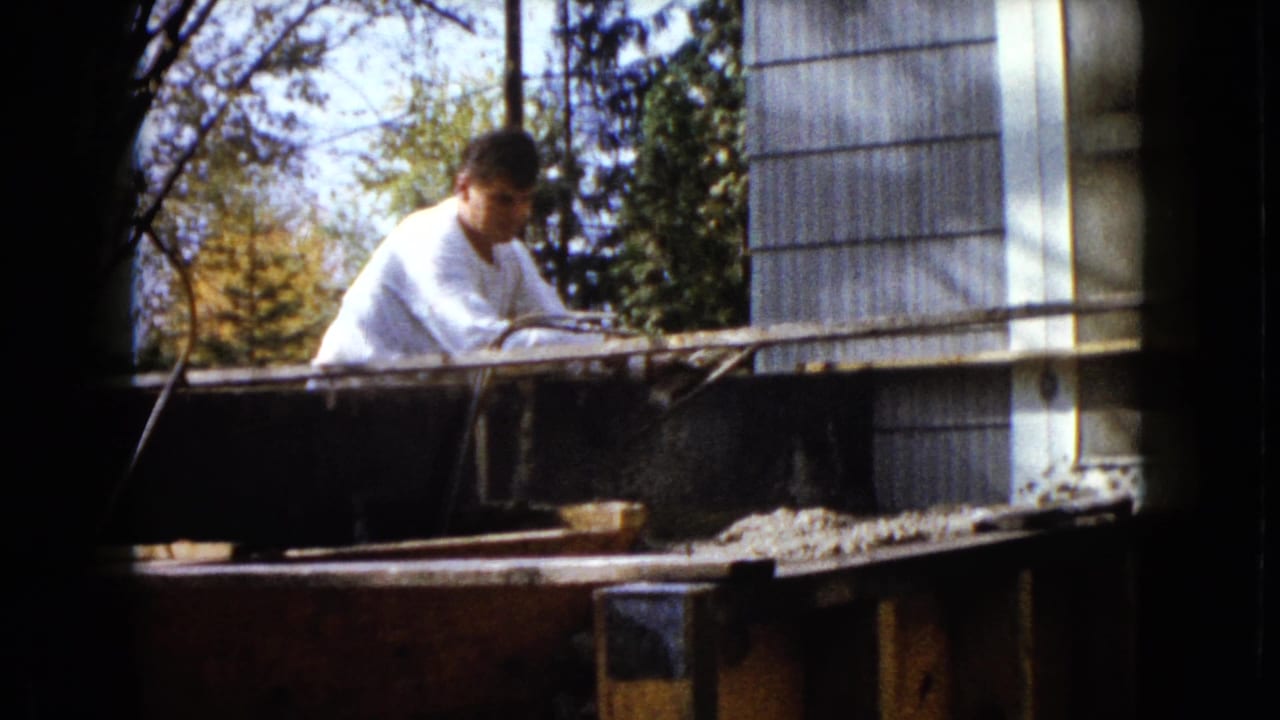 The man is more upright behind the plank, framed by house siding, a window, trees, and dark film border shadows.