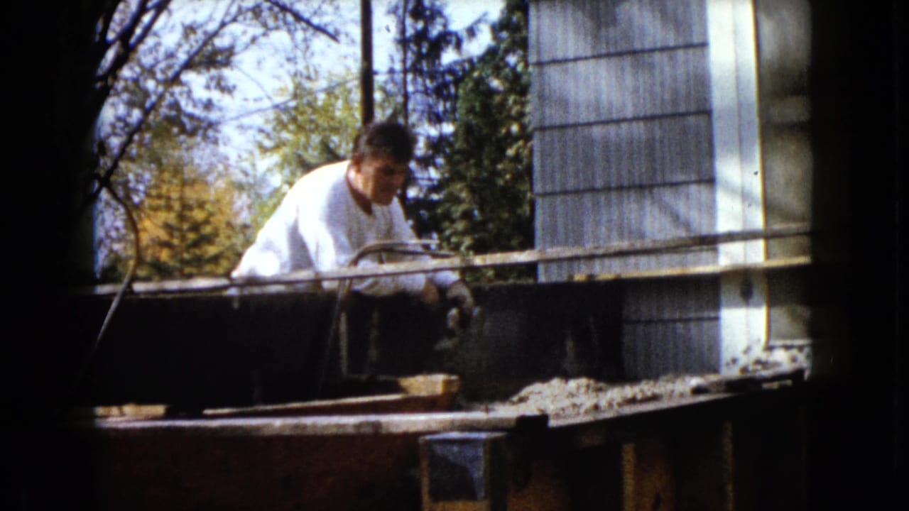 Man in a white shirt works beside a wooden porch frame near a sided house.
