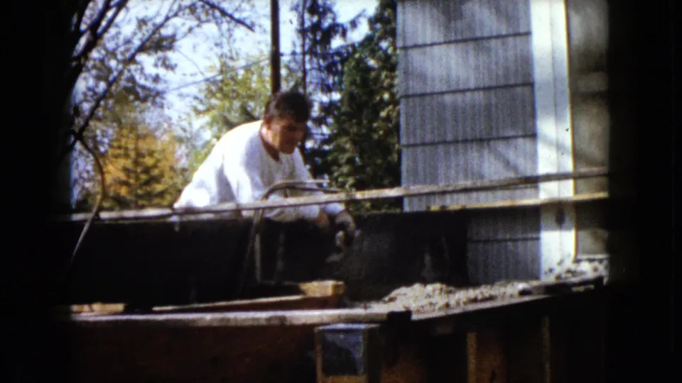 In 1960 Wauconda, Illinois, a man in a white shirt appears to work on a wooden porch frame beside a house in daylight.