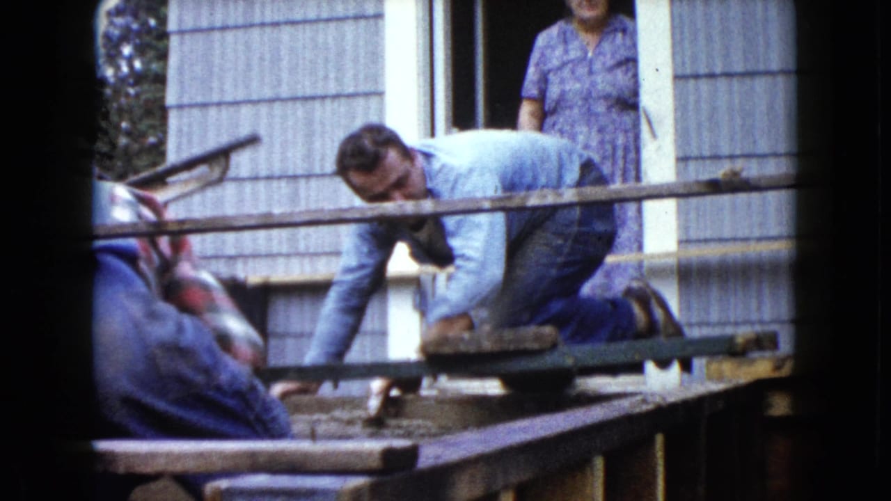 Man kneels on a porch frame while another worker and a woman in a doorway are nearby.