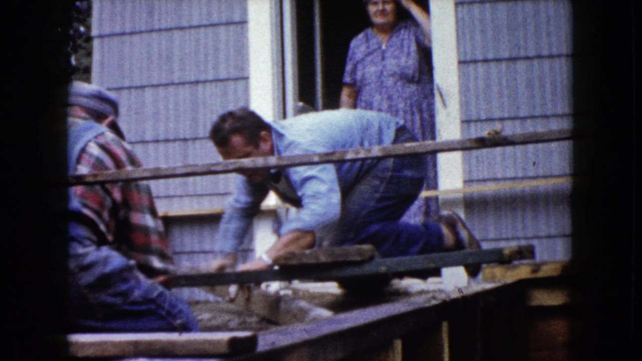 Two men continue porch construction outside a Wauconda house in 1960 while a woman in a patterned dress watches from the doorway.