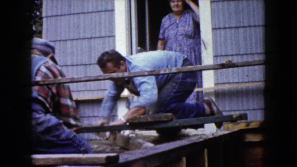 Two men continue porch construction outside a Wauconda house in 1960 while a woman in a patterned dress watches from the doorway.