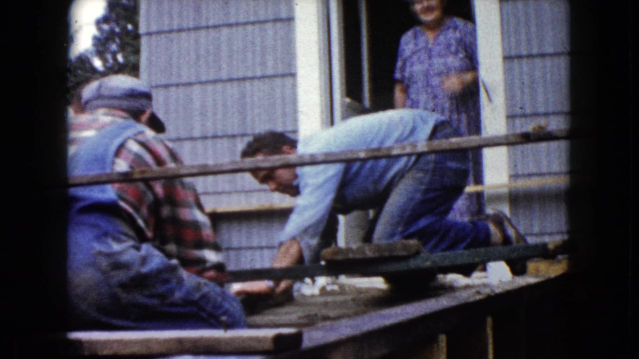 Men lean over the porch framing outside the Wauconda residence, with the doorway and watching woman still visible in the 1960 home movie frame.