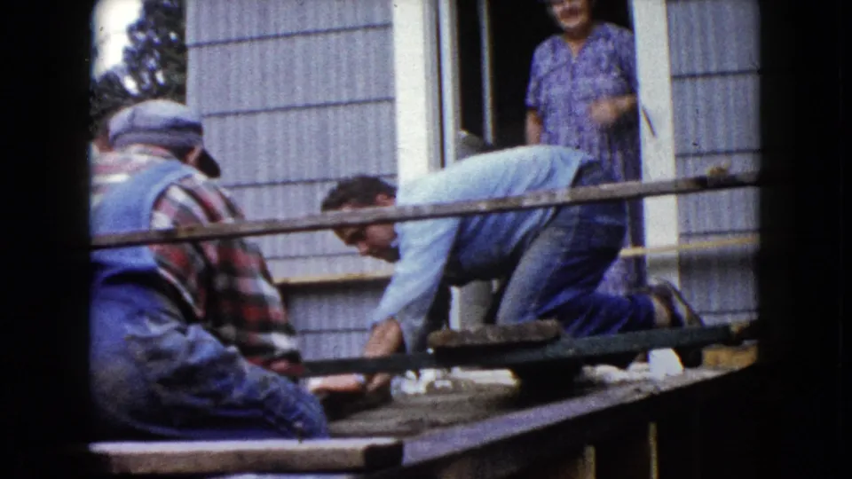 Men lean over the porch framing outside the Wauconda residence, with the doorway and watching woman still visible in the 1960 home movie frame.
