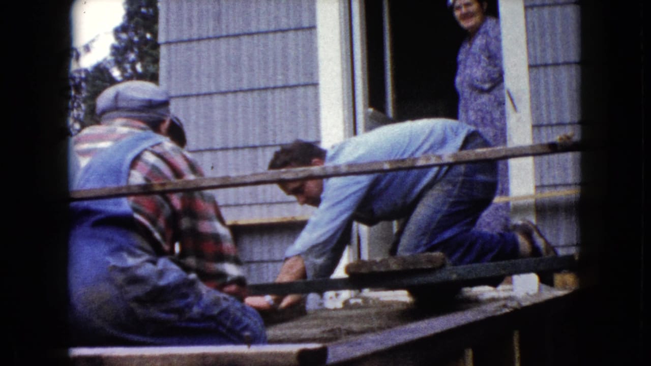 1960 Wauconda home-movie frame showing two men in work clothes on a porch structure as a woman watches from the doorway.