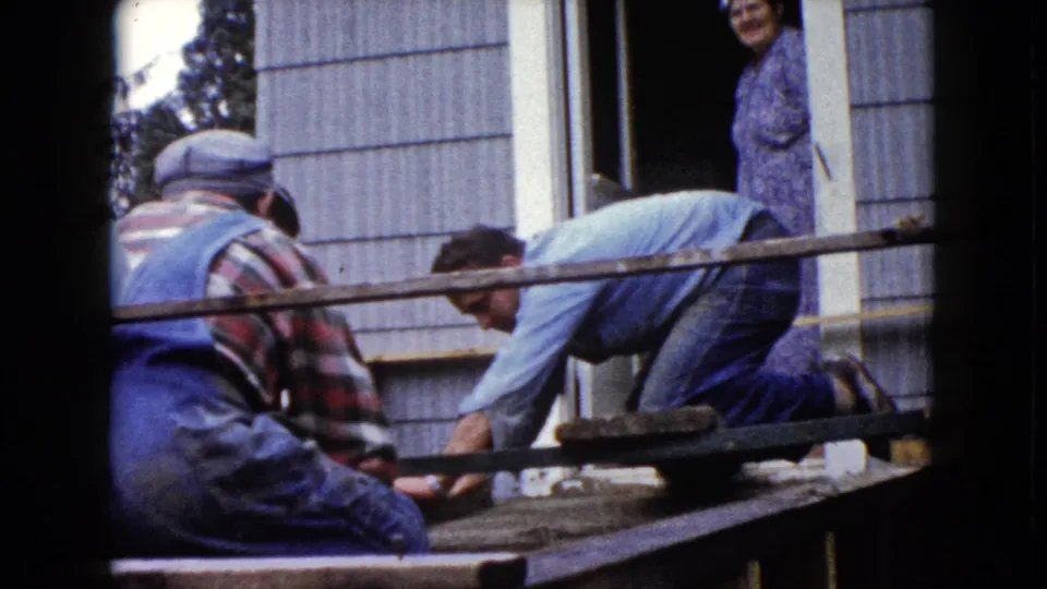 1960 Wauconda home-movie frame showing two men in work clothes on a porch structure as a woman watches from the doorway.