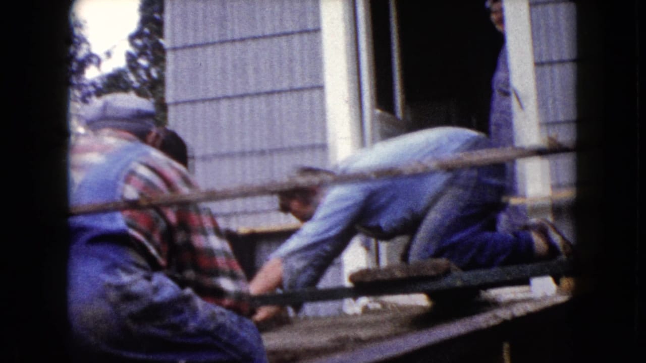 1960 Wauconda exterior frame with two men working close to a house doorway on a wooden porch or scaffold structure.