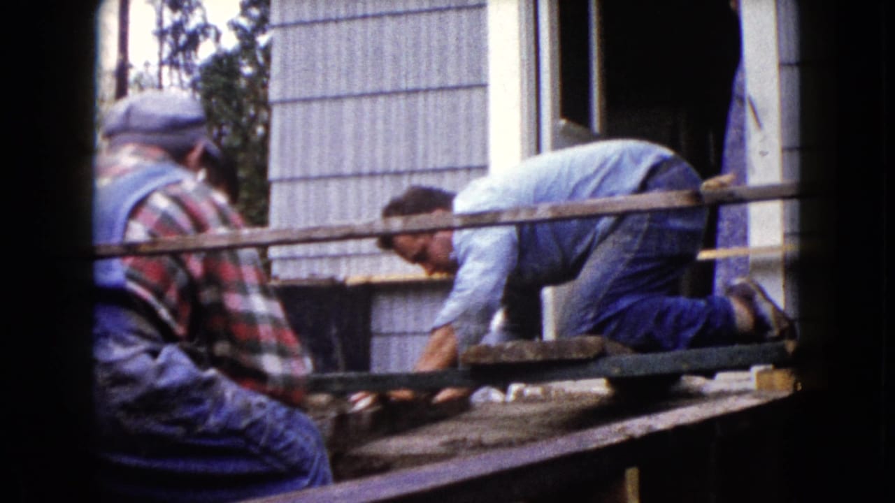 Home-movie frame from Wauconda, Illinois, showing two men focused on porch construction or repair in daylight.