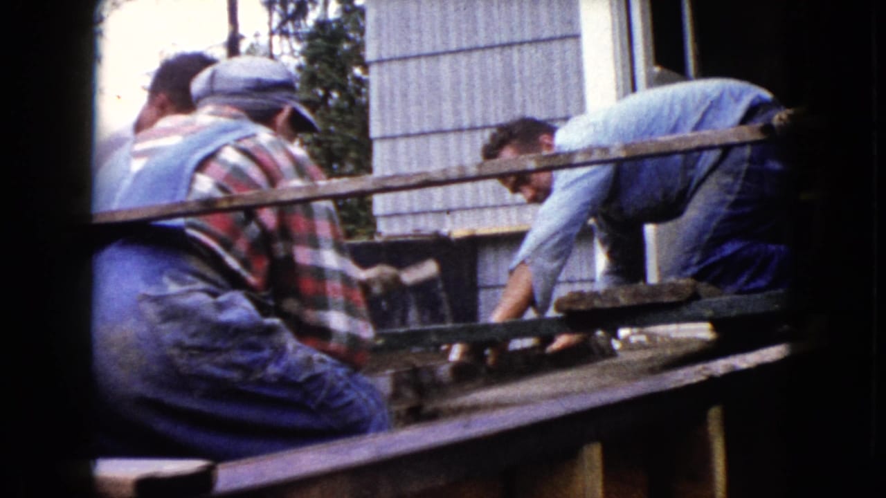 1960 Wauconda home-movie frame showing men in work clothes leaning over a porch area with wet concrete and tools.