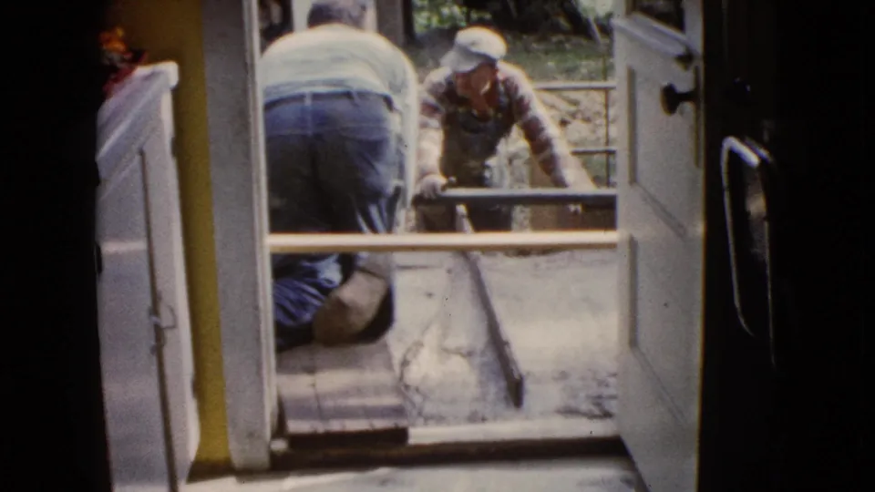 1960 Wauconda 8mm frame with a kneeling worker inside the doorway and another man handling porch concrete outside.