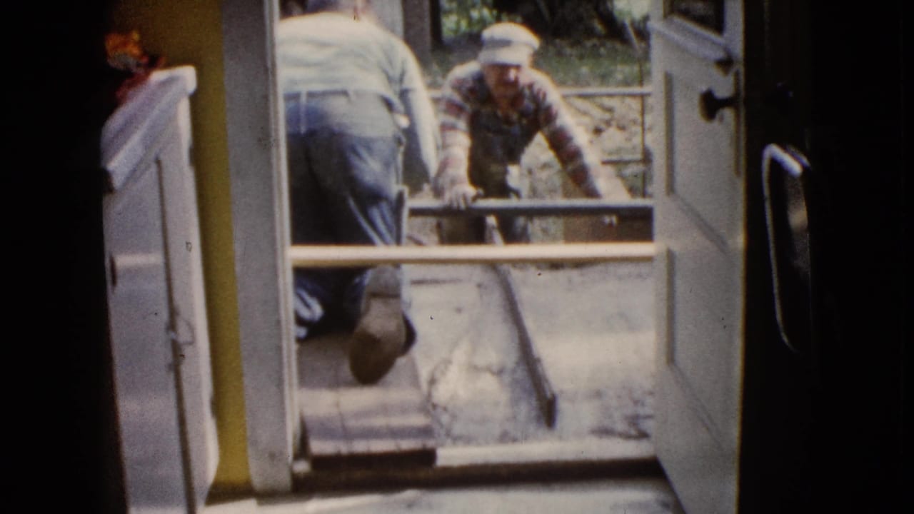 1960 Wauconda home-movie frame showing men at an open doorway during porch concrete work.