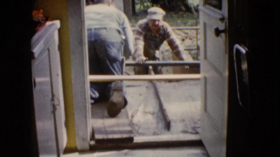 1960 Wauconda home-movie frame showing men at an open doorway during porch concrete work.