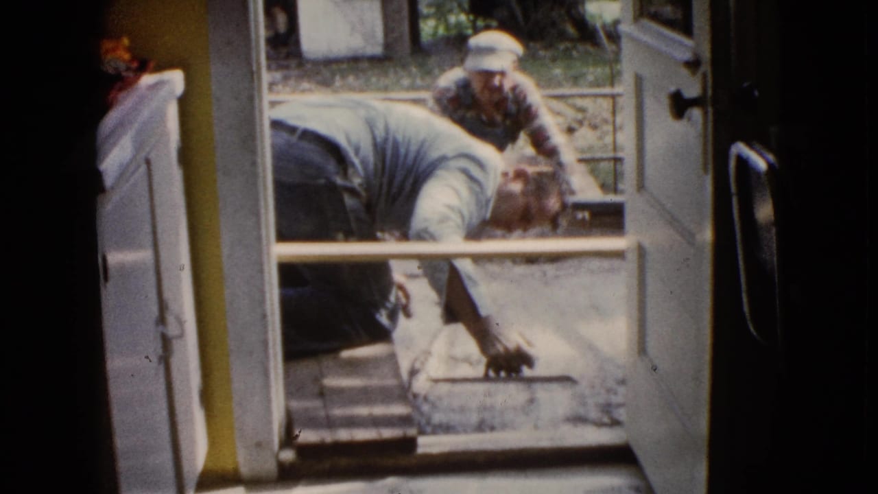 A man in a light shirt reaches down with a tool as another man faces the doorway during the 1960 cement work scene.