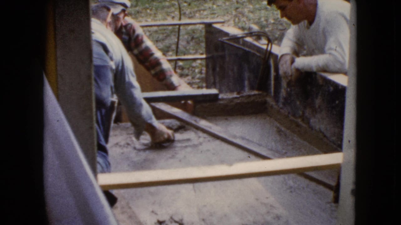Three older men work around wet cement in a rectangular form viewed through a doorway.