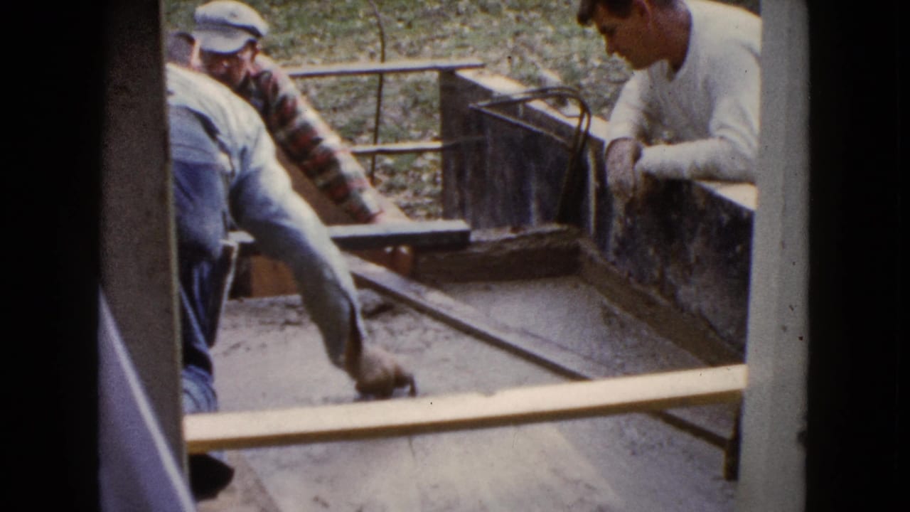 A man bends over wet cement with a screed while two men watch from nearby.