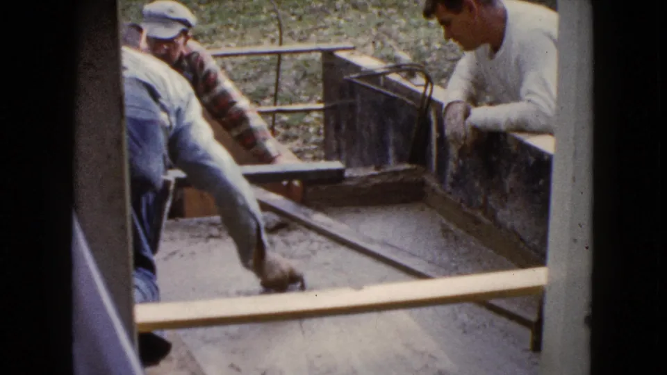 A 1960 Wauconda home-movie frame shows one man leveling cement with a board as two older men look on.