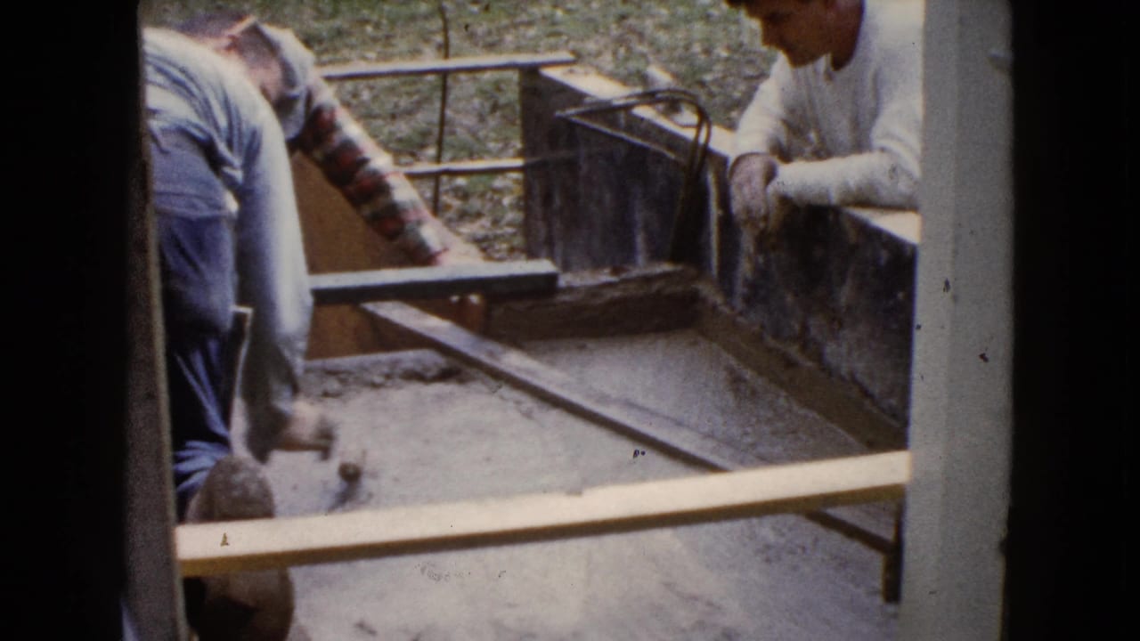 Two men in 1960 Wauconda construction footage work beside wooden form boards, with wet concrete and a leveling tool in view.
