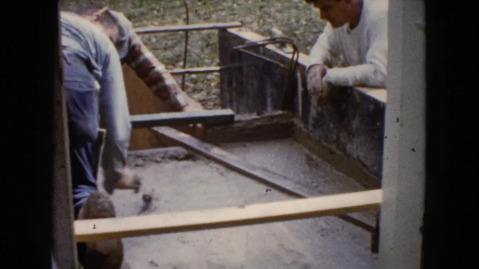 Two men in 1960 Wauconda construction footage work beside wooden form boards, with wet concrete and a leveling tool in view.