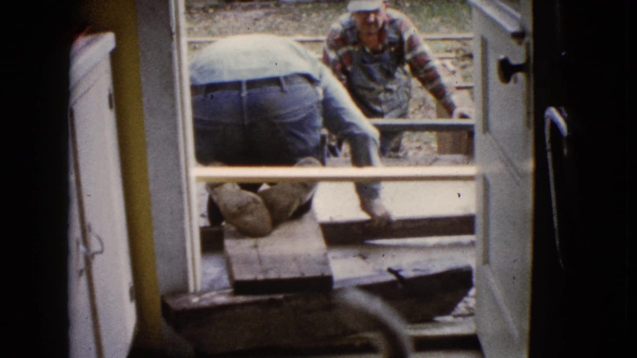 The 8mm frame shows a kneeling worker at the concrete foundation and a second man beyond the form, seen through an open doorway.