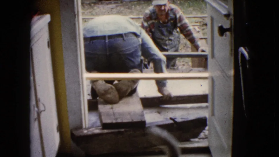 The 8mm frame shows a kneeling worker at the concrete foundation and a second man beyond the form, seen through an open doorway.