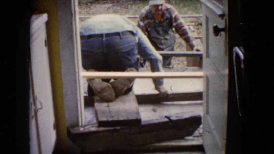 1960 Wauconda home-movie frame showing men repairing a porch or patio surface through a doorway.