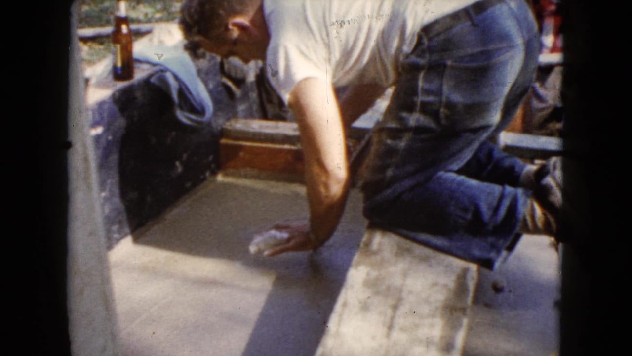 Man in white T-shirt kneels over wet cement with a trowel nearby.