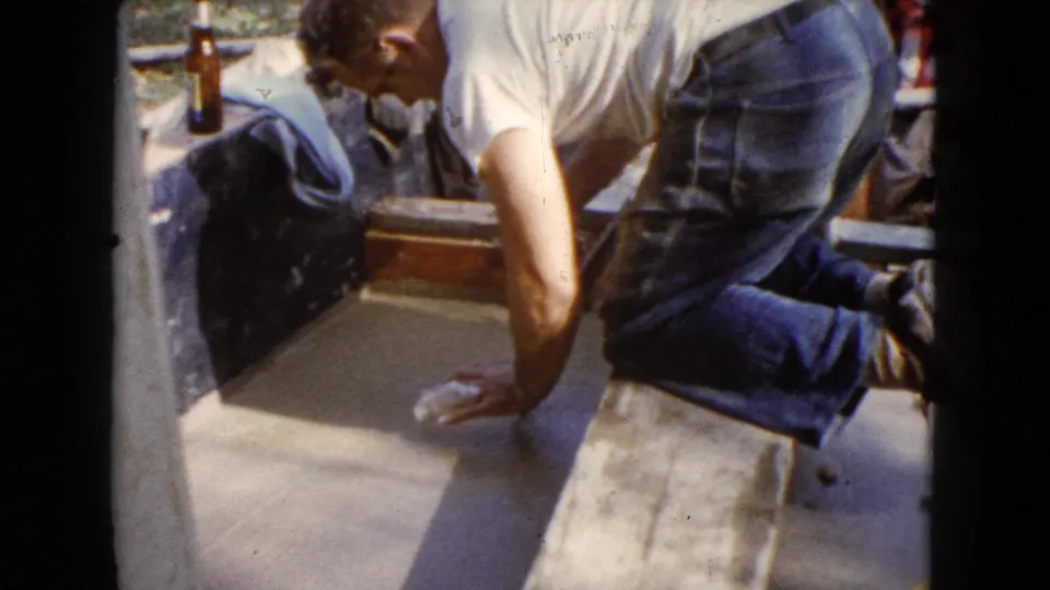 A man in a white T-shirt and jeans kneels beside wet cement during a 1960 home repair in Wauconda, Illinois.